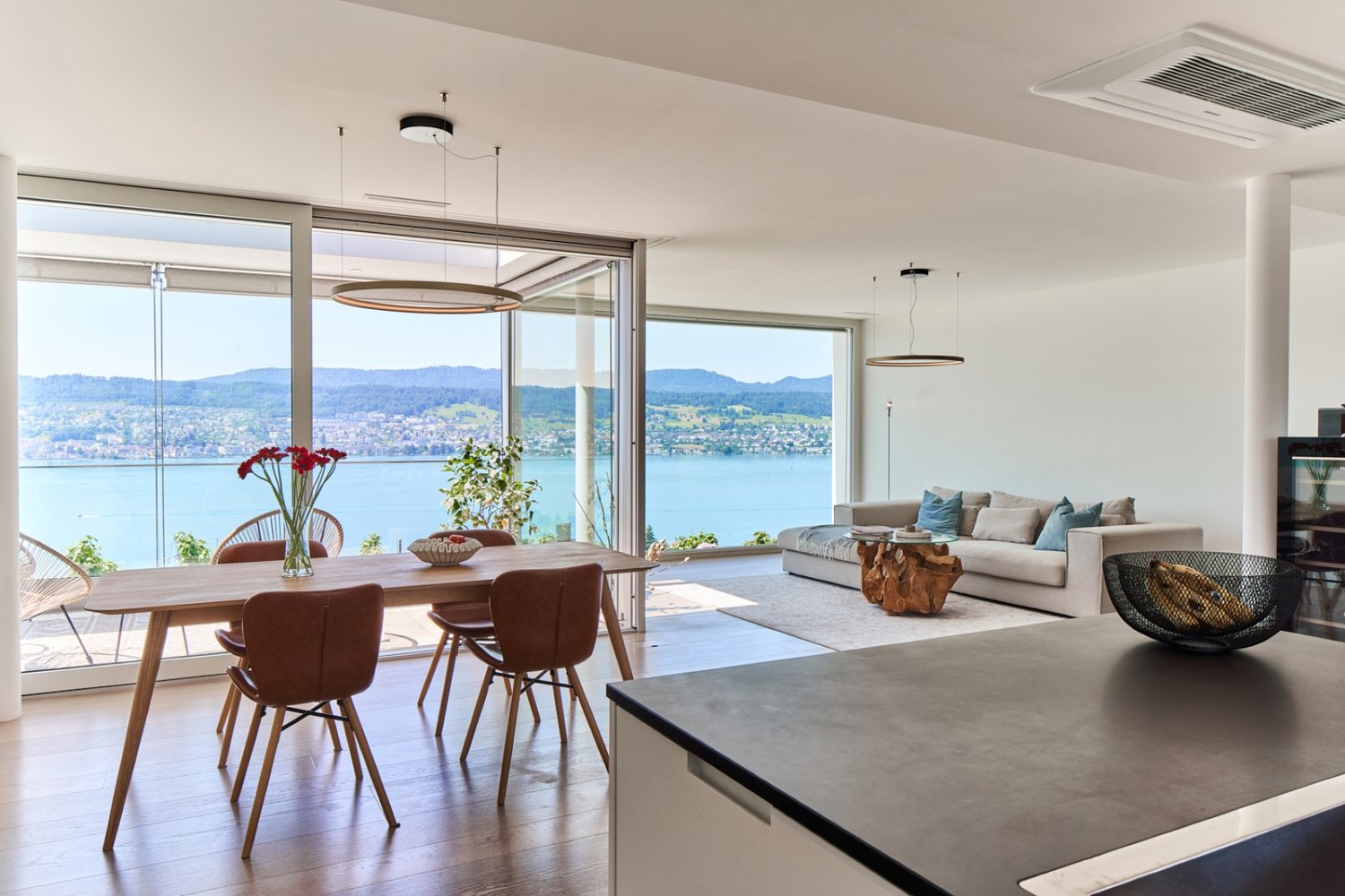 Spacious open-plan living and dining area with large windows offering a scenic view of the lake and surrounding mountains. The dining table with wooden chairs is situated near the windows, while the living room features a comfortable sofa and armchairs. T
