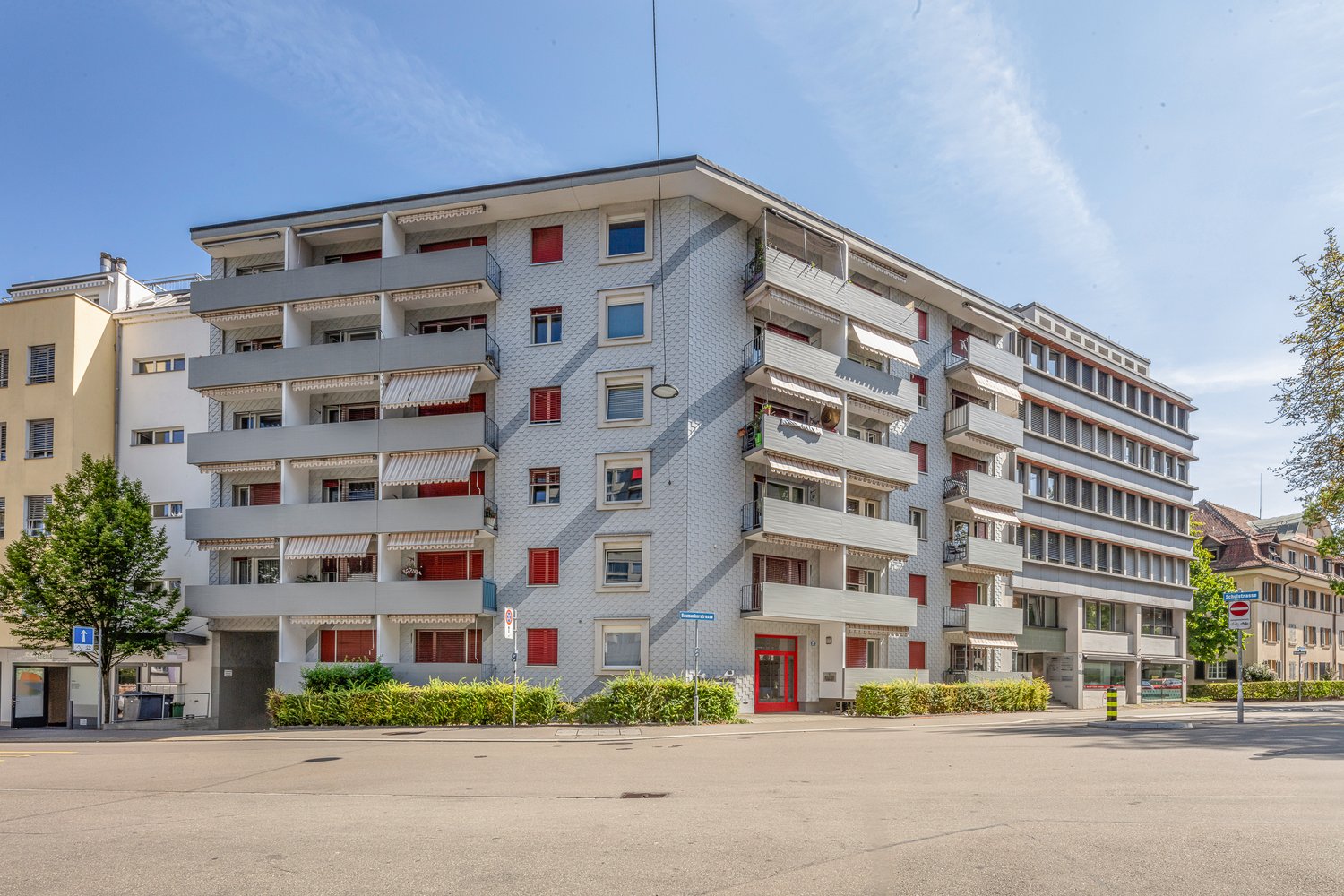 An apartment building with a white brick wall and a red door on the first floor, a few balconies on each floor, and a red shutter for windows, trees, and bushes in front of the building