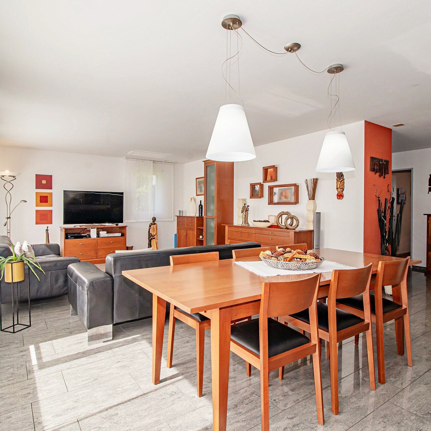 Dining room with a wooden dining table, 4 wooden chairs, a bowl of snacks, gray sofa, black leather footrest, wooden entertainment center, television, indoor plants, lamps, cabinets, shelves, decorative items, and artwork on the wall