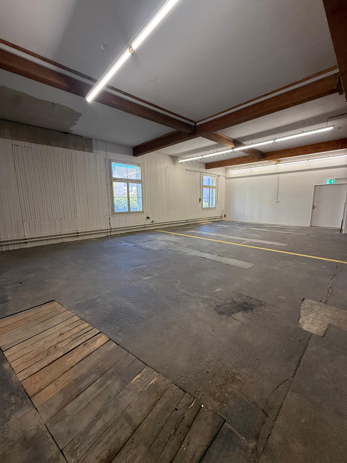 empty room with wooden planks on the left, metal beams and lights on the ceiling, windows on the left wall