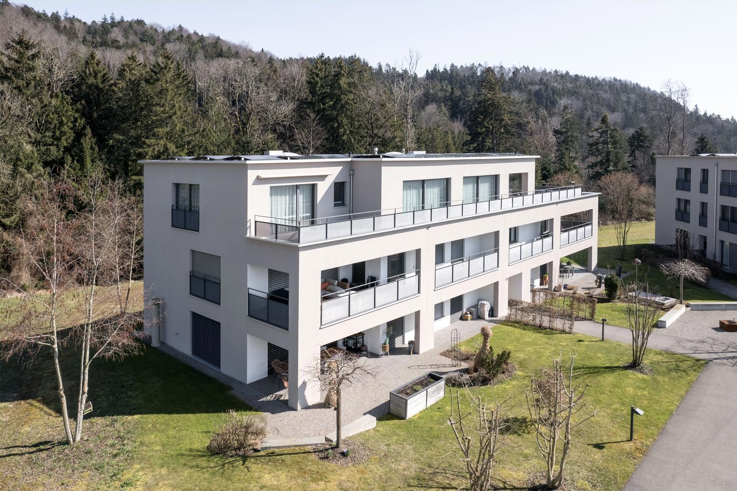Modern white building with three floors, balconies on all floors, solar panels on the roof, surrounded by trees, grassy areas and a small path