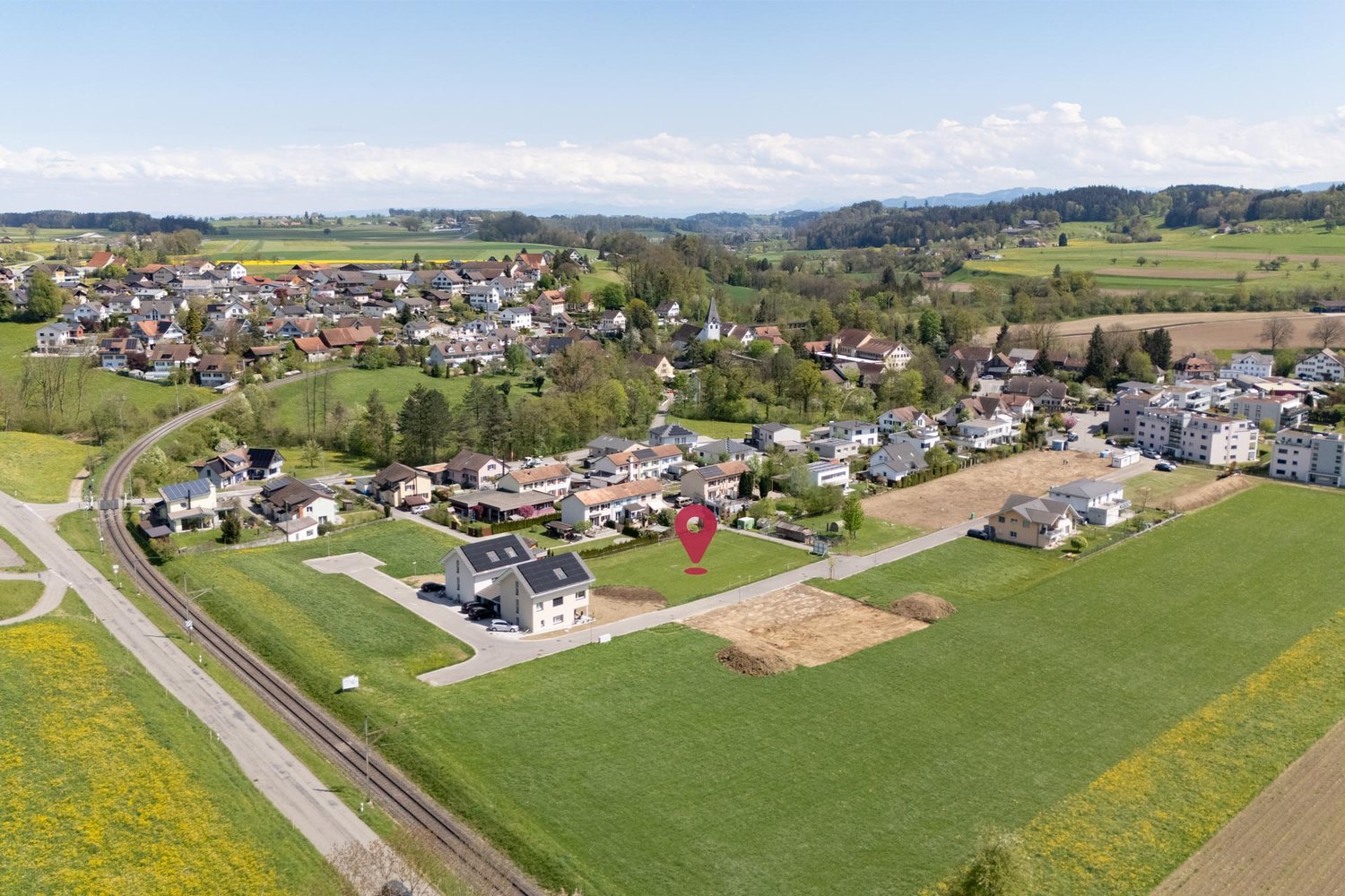 This image shows an aerial view of a small town or village surrounded by rolling hills, fields, and forests. The town has a mix of residential and commercial buildings, with a church steeple visible in the center. There is a road running through the town,