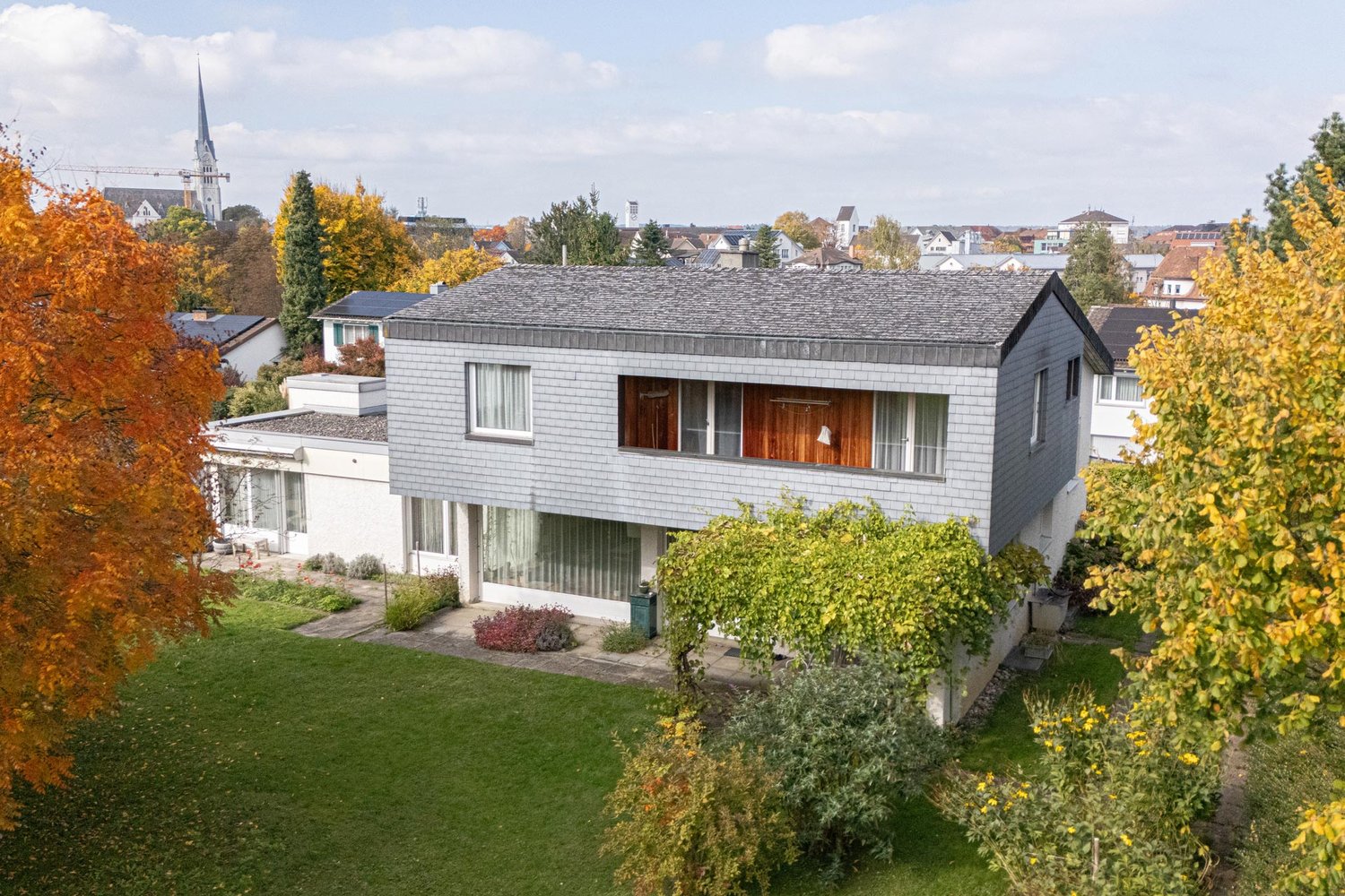 Modern two-story house with a flat roof, white walls, glass windows, wooden doors, solar panels on the roof, surrounded by green grass and trees.