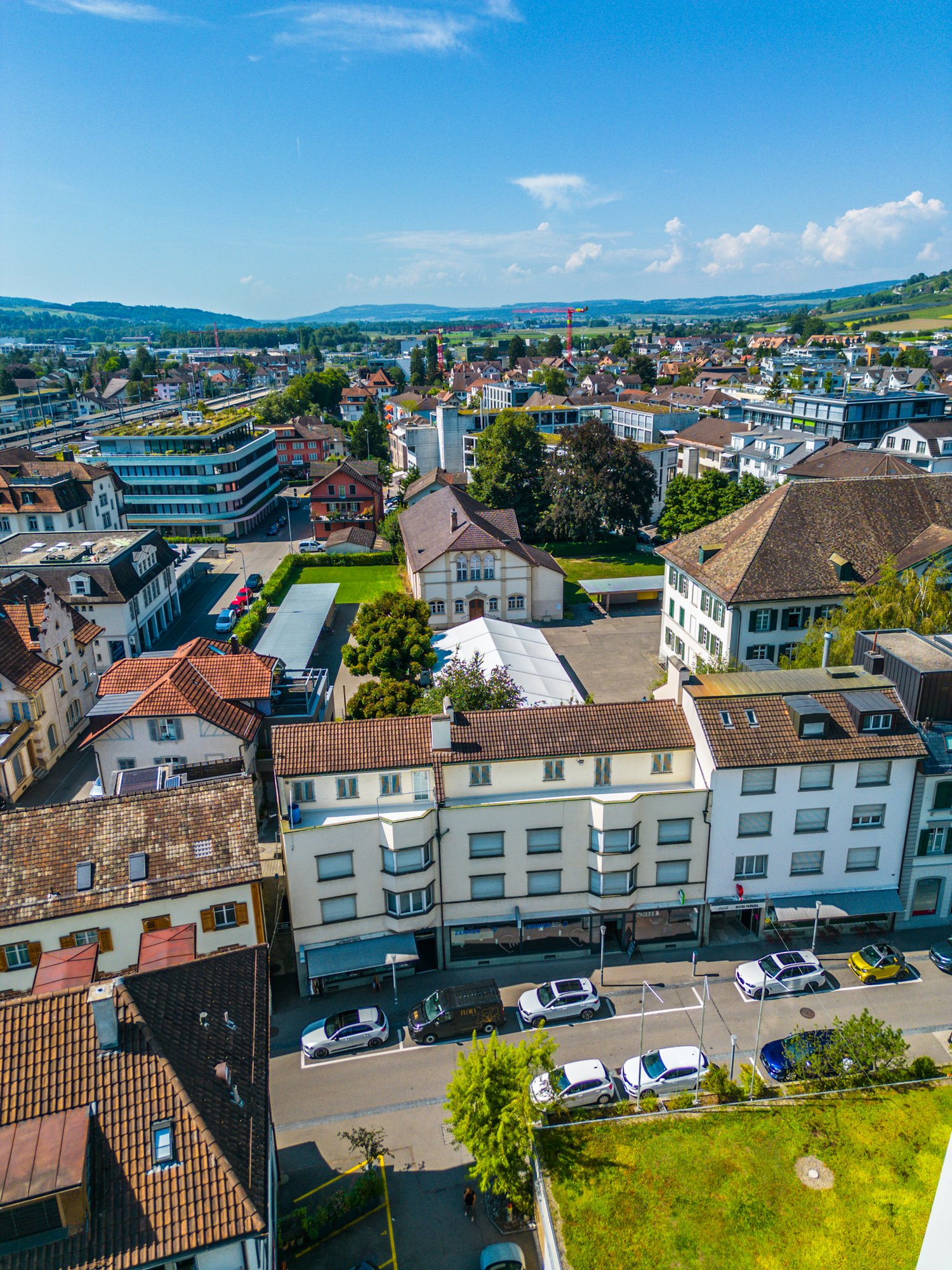 Cityscape with multiple buildings, some with cars parked in front, green spaces and trees in between