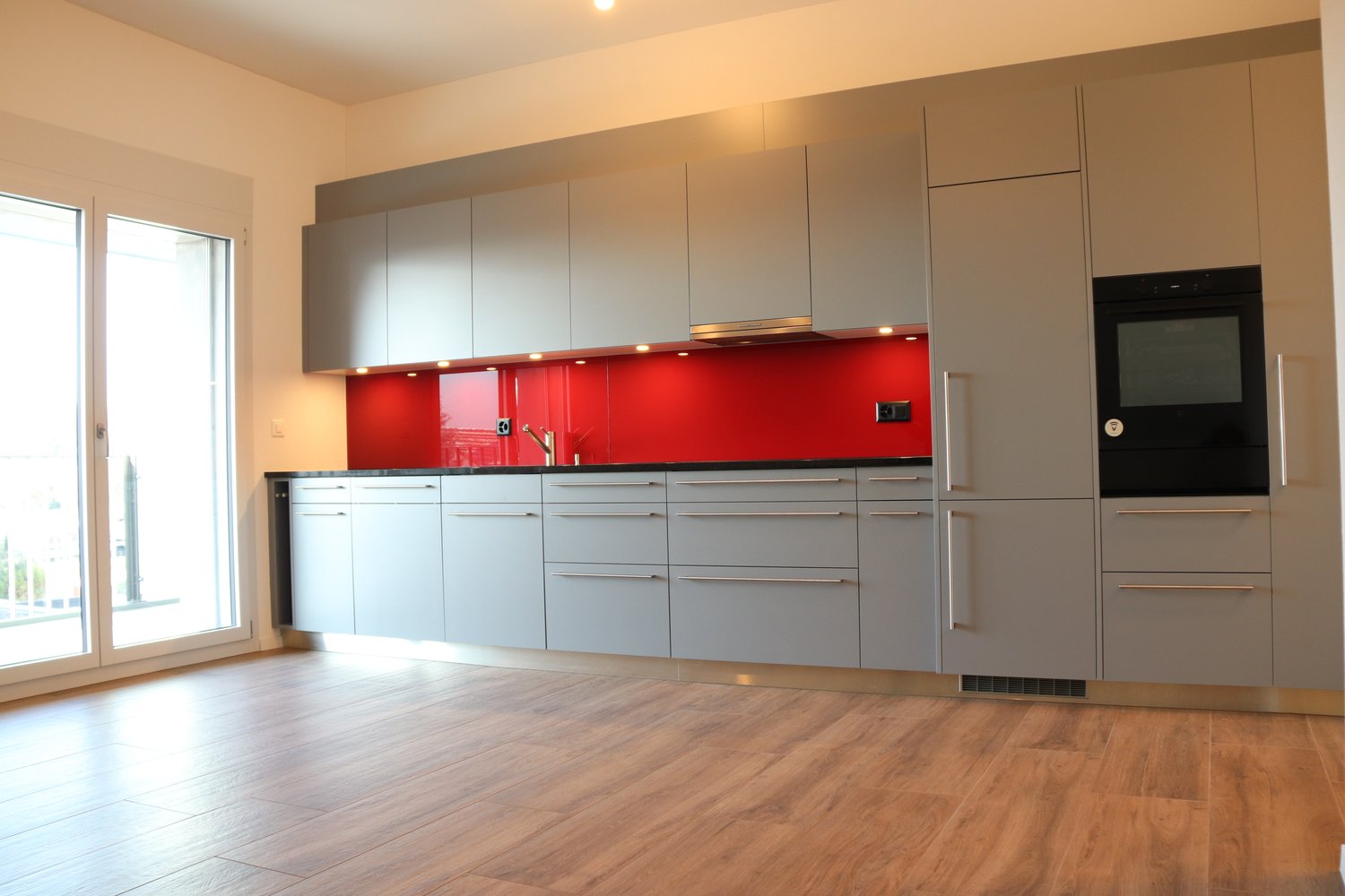 Modern kitchen with gray cabinets, red backsplash, stainless steel faucet, black oven, and wooden floors.
