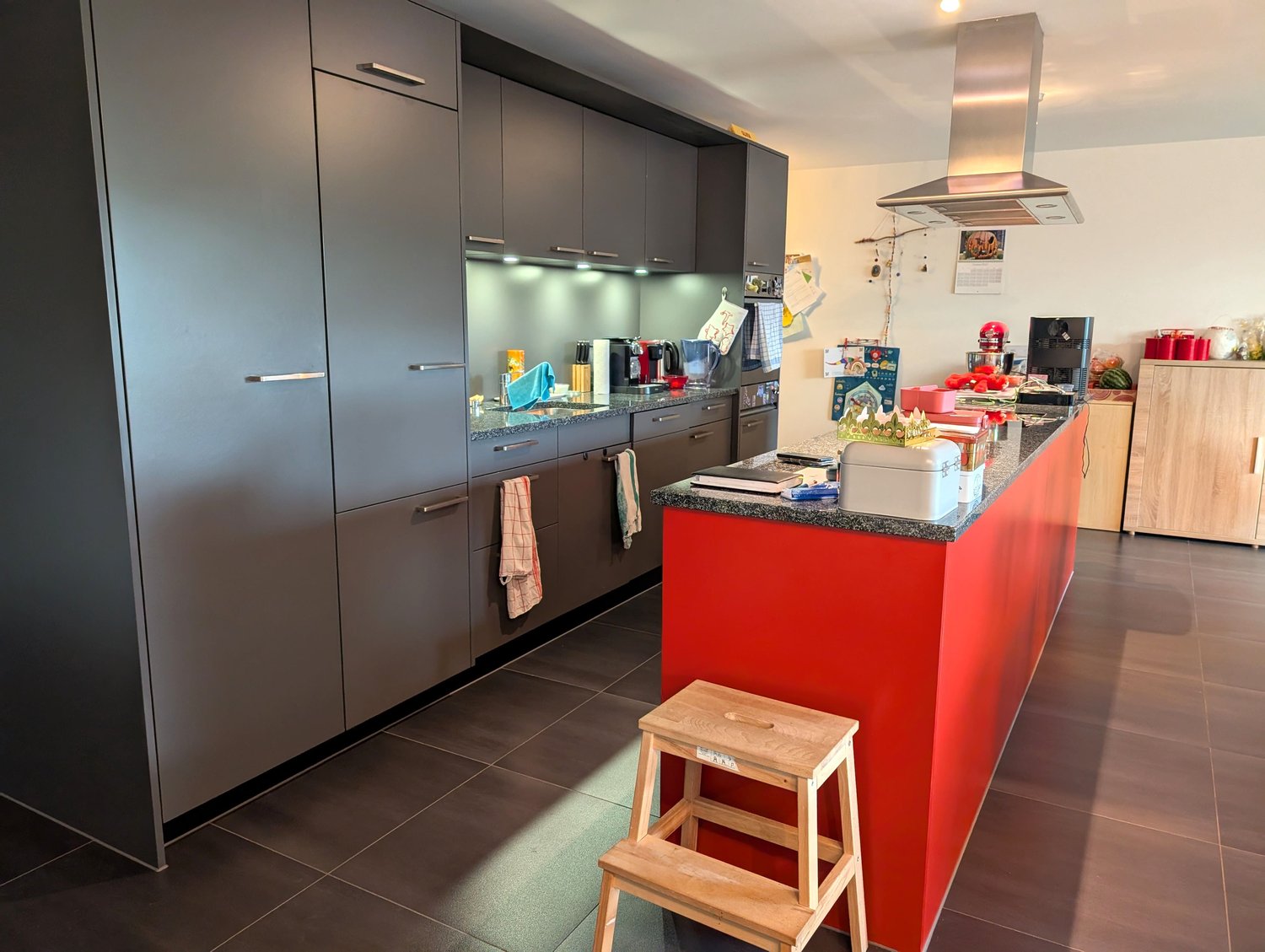 Modern kitchen, gray cabinets, red island counter, wooden step stool, wooden cabinets on the right side, stainless steel range hood, tiled floor.