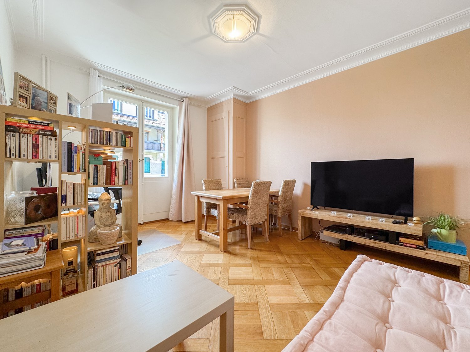 open living room with wooden floor, white walls, shelves filled with books, a TV, a table, chairs, a dining table, a Buddha statue, a window with curtains, a potted plant, a couch
