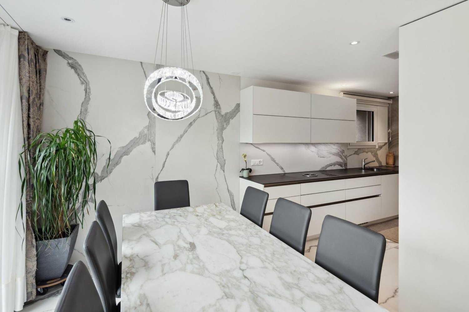 The image shows a modern and stylish kitchen and dining area. The kitchen features white cabinets and a marble-like countertop. A dishwasher is visible in the kitchen. The dining area has a large marble-top table with black leather chairs. The room is dec