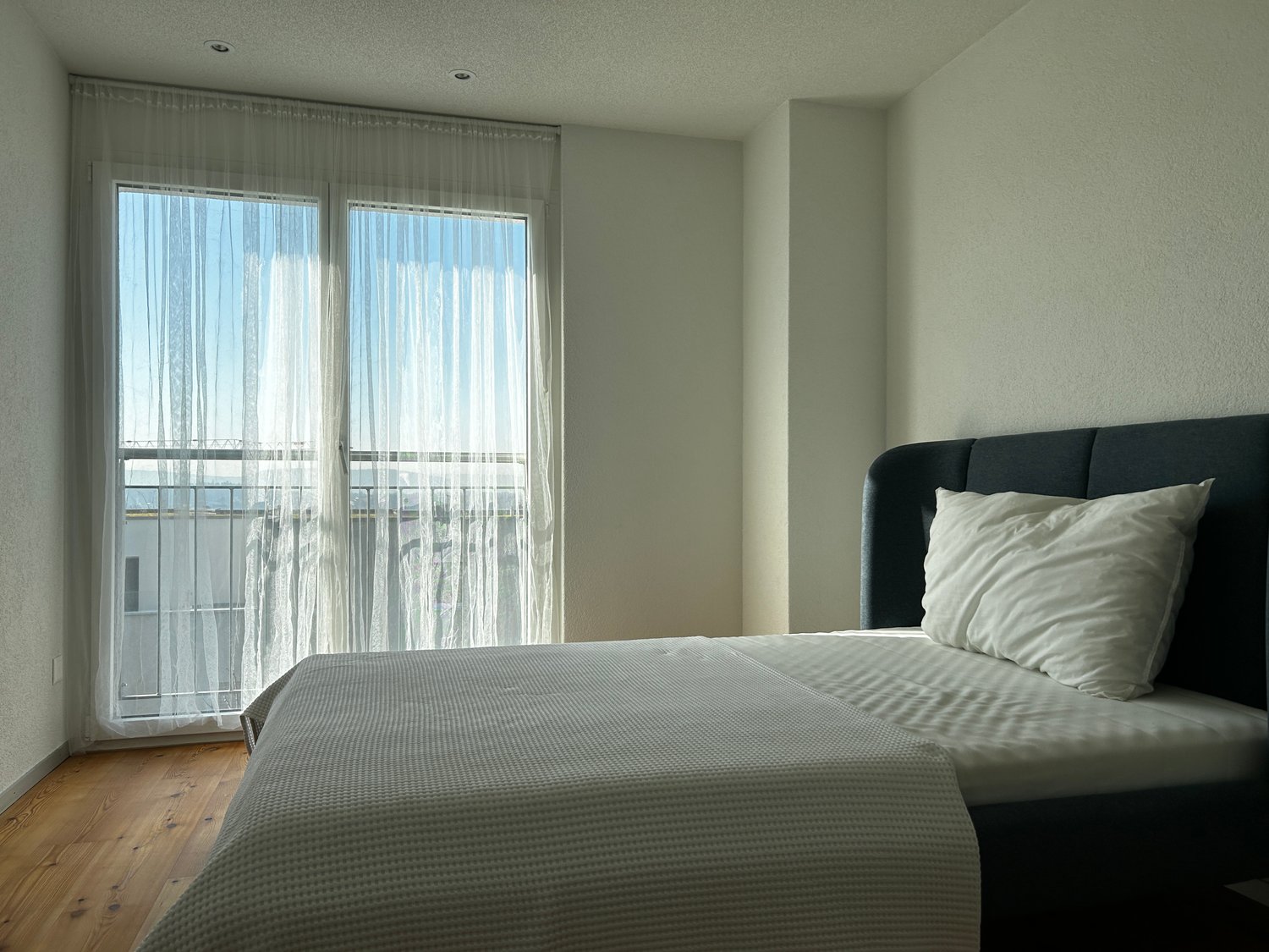 Large bedroom with a black upholstered bed frame, white bedding, and large windows with sheer curtains overlooking a balcony or terrace.