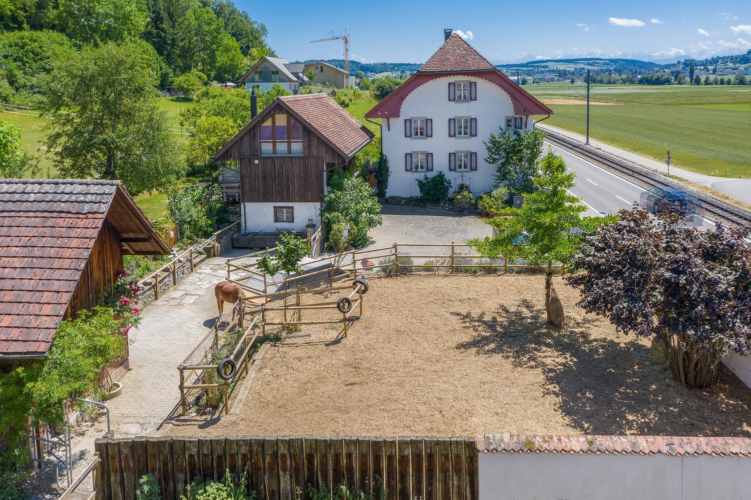 Two-story traditional Swiss-style house with a wooden barn-like structure, surrounded by a fenced yard with a horse, located in a rural setting with fields and mountains in the background