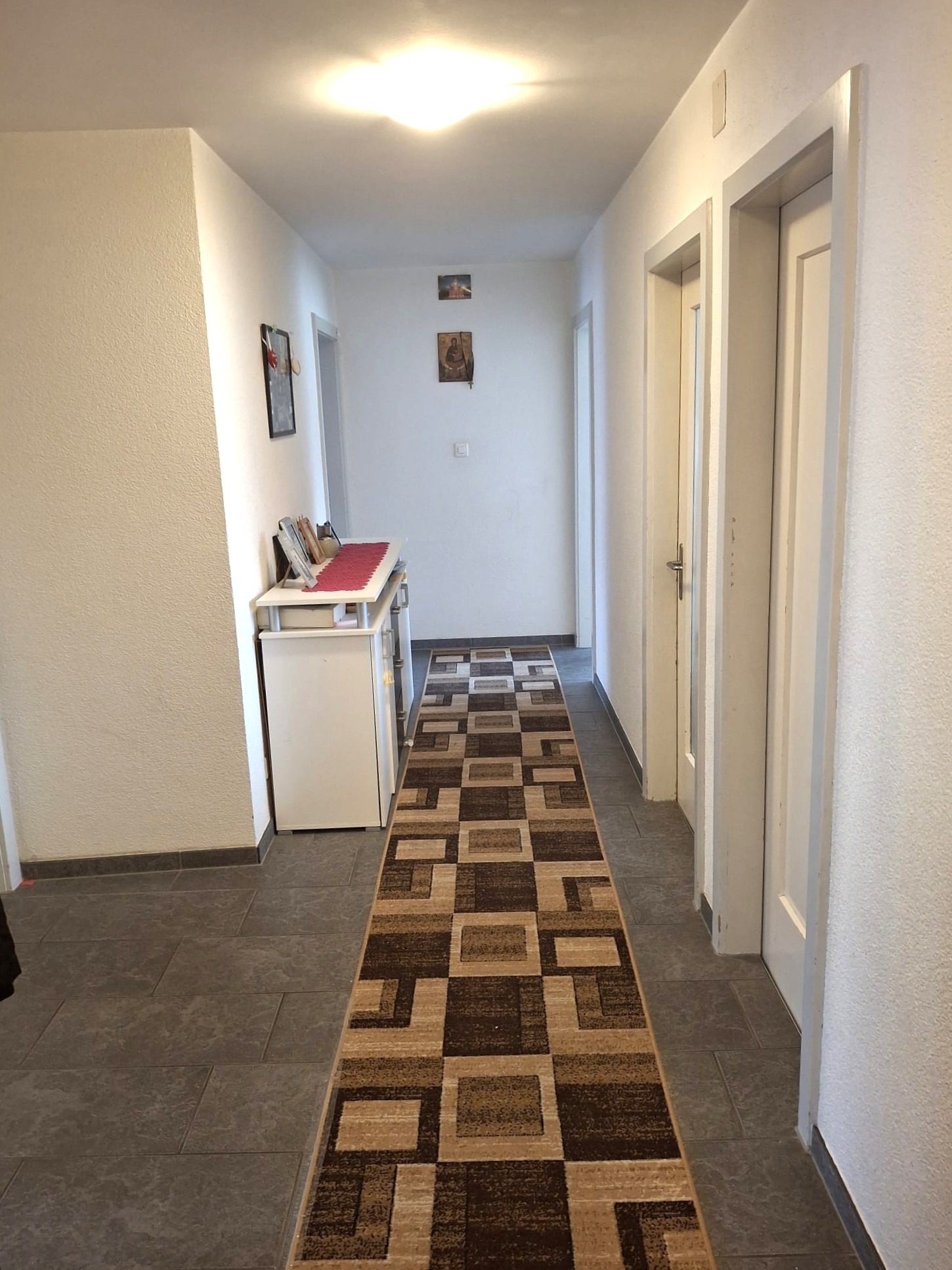 The image shows a hallway with white walls, a tiled floor with a patterned carpet runner, and a small table with some items on it. There are two doors visible, likely leading to other rooms. The lighting appears to be a ceiling-mounted light fixture.