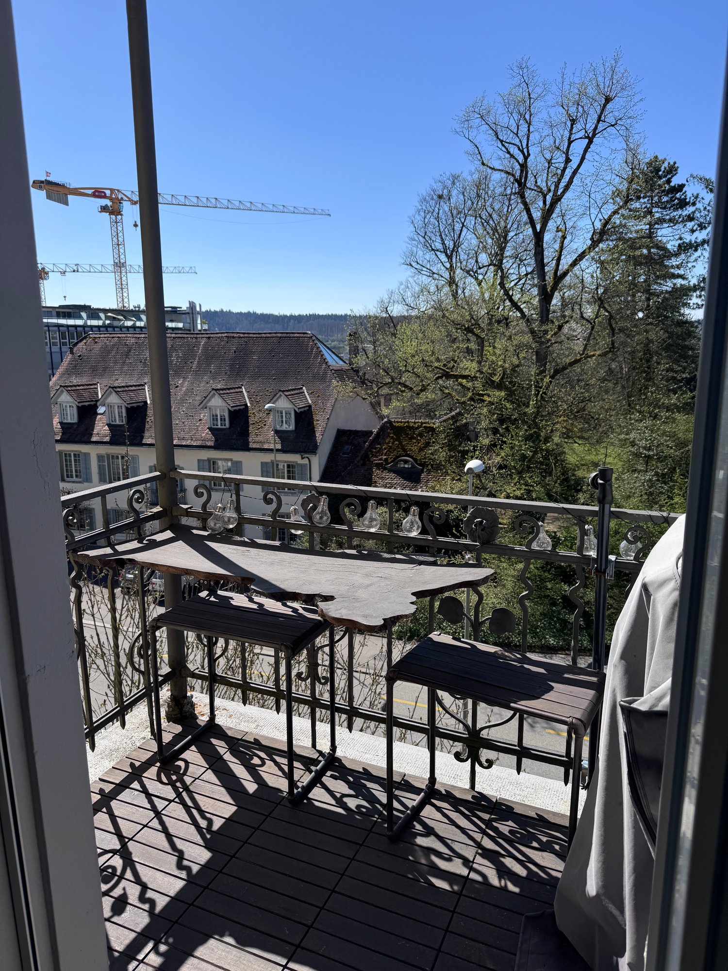 A balcony with a wrought iron railing and a small table with chairs, overlooking a view of rooftops and trees in the distance. A construction crane is visible in the background.