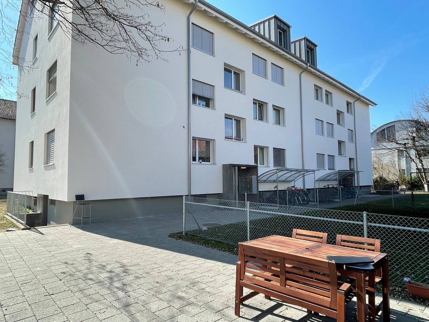 4-story apartment building with white exterior, large windows, and a covered entrance. There is a paved courtyard area with a wooden table and chairs, as well as a fenced-in area that appears to be used for parking.
