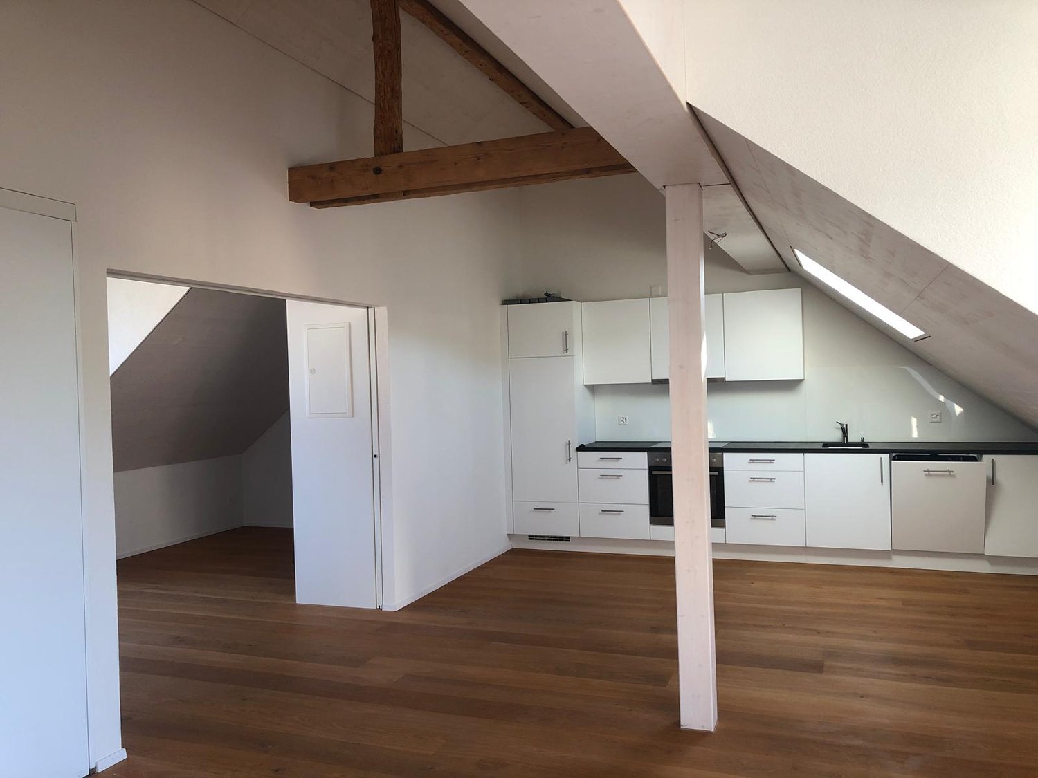 Open-concept kitchen and living area with hardwood floors, white cabinets, and a dishwasher. Exposed wooden beams on the ceiling add a rustic touch. The space has a modern and minimalist design.