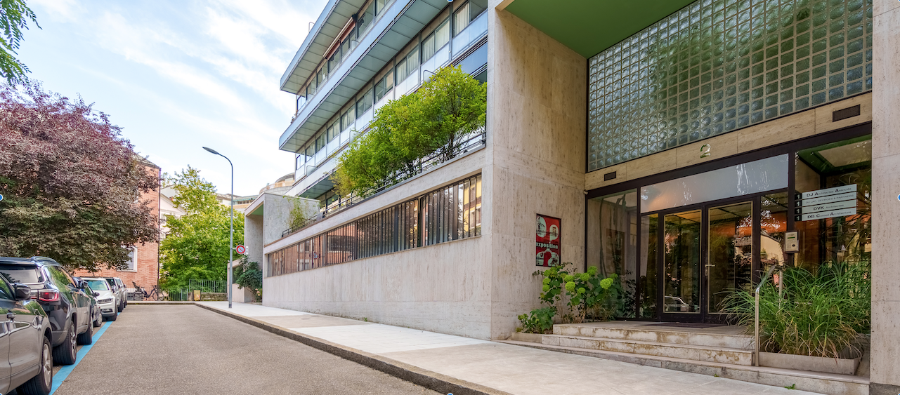 Multi-story building with concrete and glass facade, balconies, and trees surrounding the entrance. The building appears to be a commercial or office space with a storefront on the ground level.