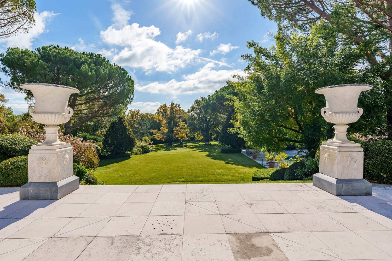 large open green lawn with trees, plants, marble vases, bright blue sky, sunny
