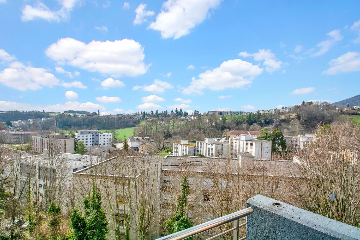 The image shows a panoramic view of a city surrounded by hills and trees. The city skyline is visible, with a mix of residential and commercial buildings. The sky is blue with scattered white clouds, creating a pleasant and scenic atmosphere.
