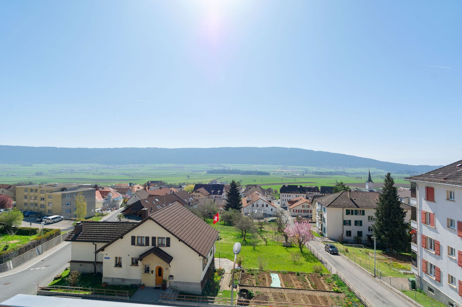 The image shows a scenic view of a small town or village nestled in a valley surrounded by rolling hills and mountains. The town has a mix of residential and commercial buildings with red-tiled roofs, and there are trees and greenery throughout the landsc
