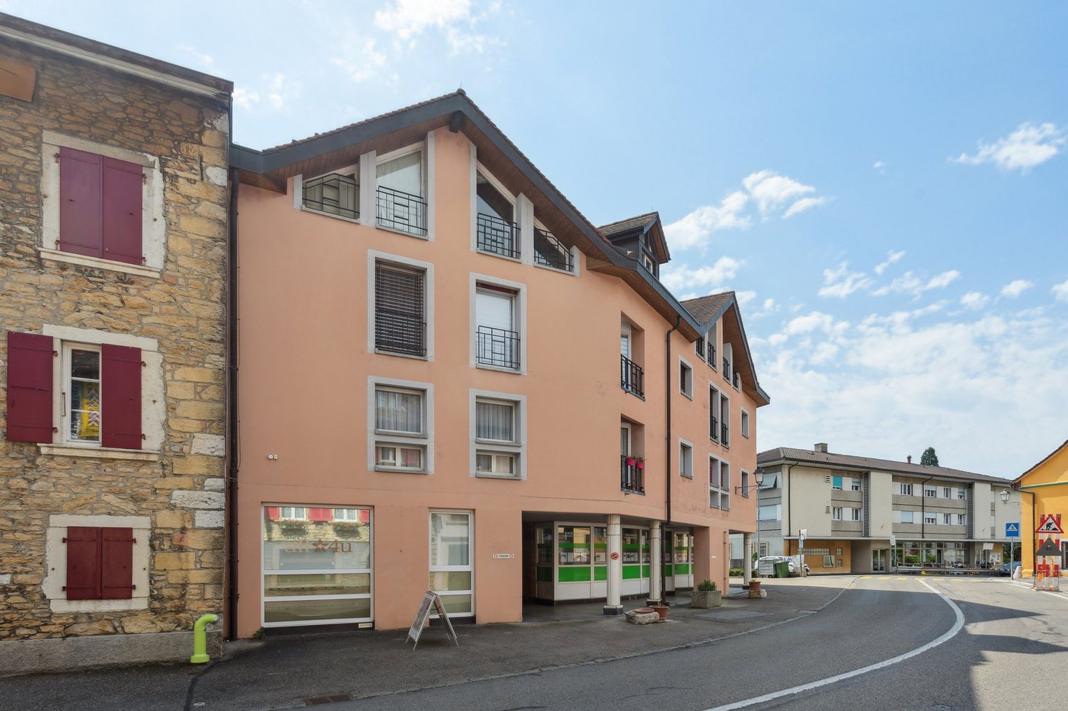 An apartment building with multiple windows, brick wall, red shutters, an entrance with a glass door, a paved road, and a green plant in front.