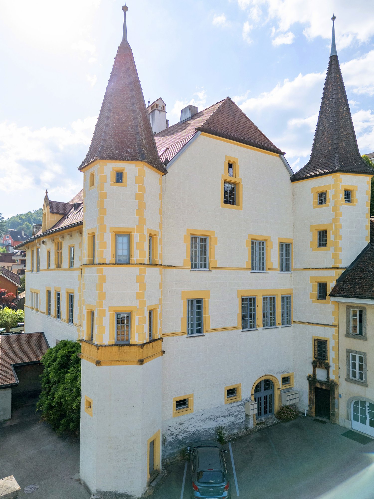 Large building with yellow trims, two spires, multiple windows, brown roofs, car parked in front