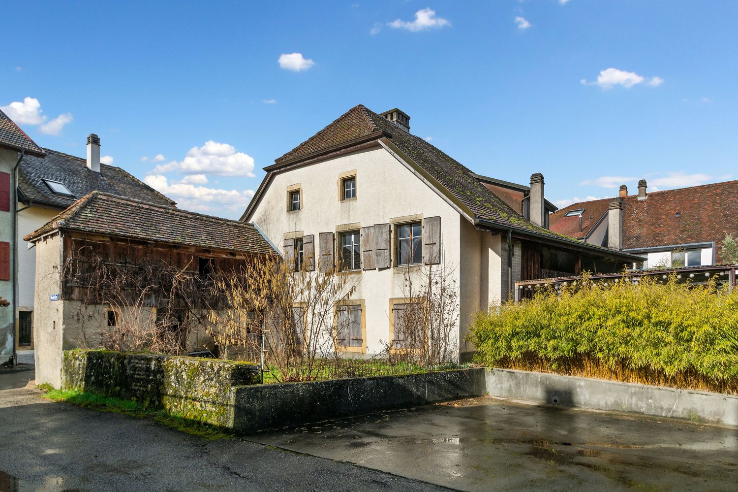 Old house, white walls, brown roof, multiple windows, chimneys, small garden, concrete wall, bushes