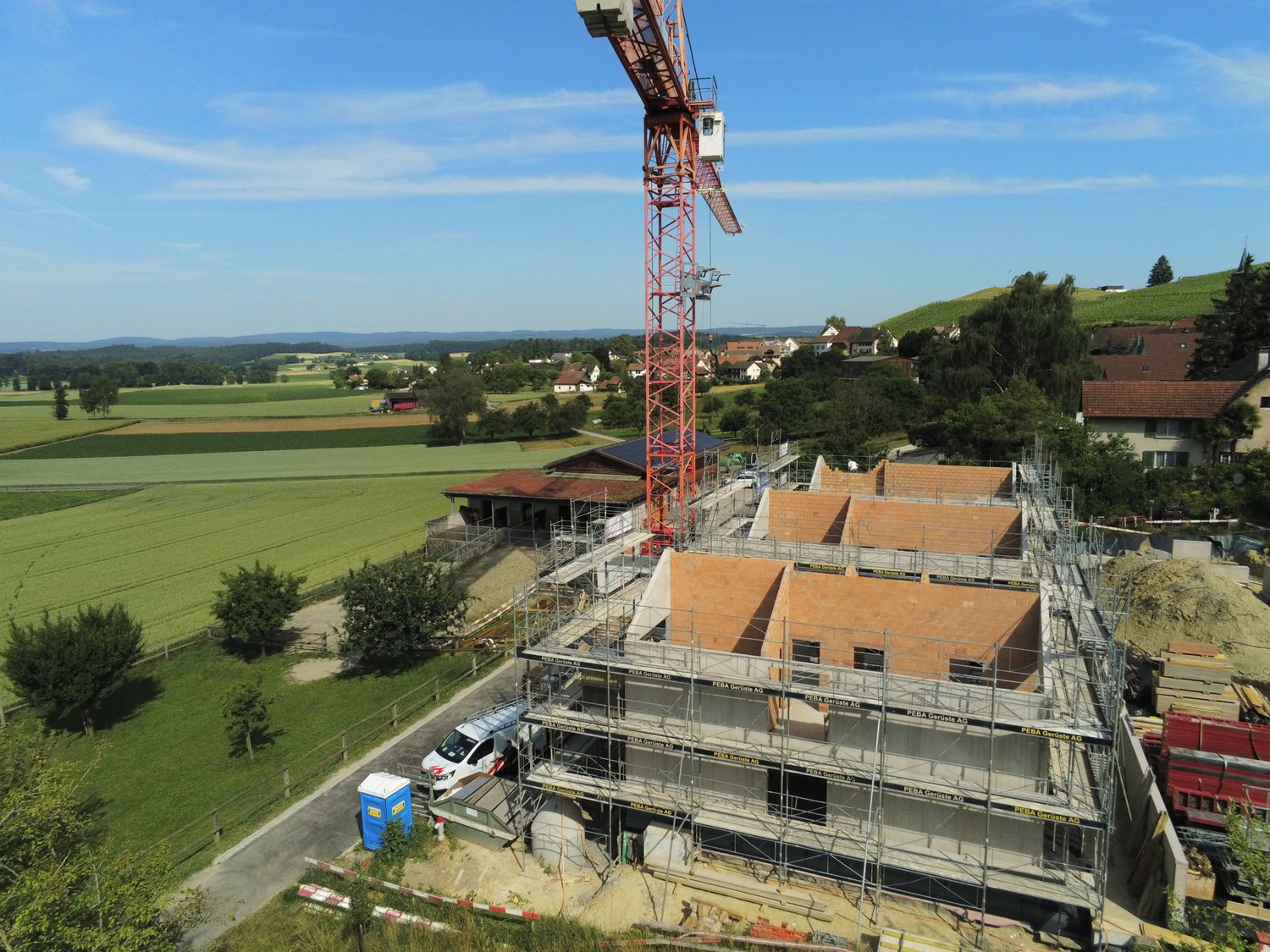 The image shows a construction site in a rural area. There is a large crane tower and scaffolding surrounding a partially constructed building. The surrounding landscape includes green fields, trees, and residential buildings.