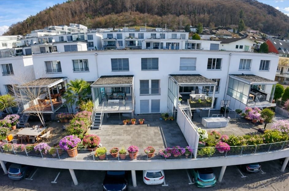 White multi-story building with multiple balconies, plants, flowers, cars parked in the ground floor parking, tables and chairs outside