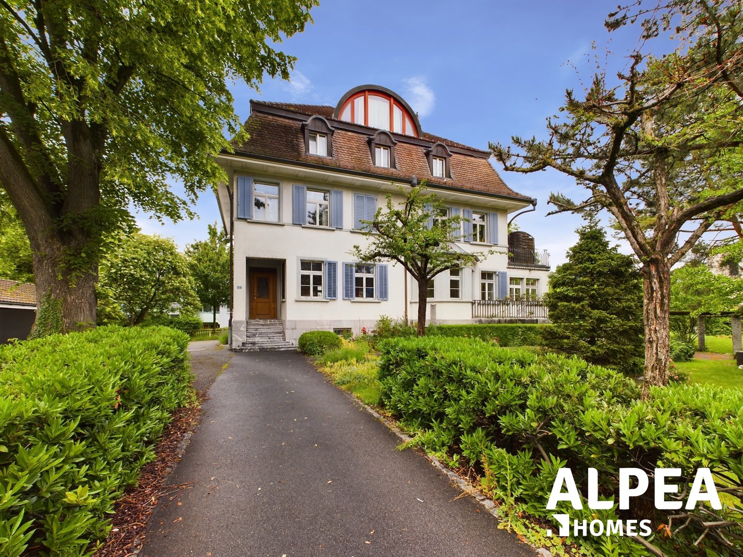 Large house, white exterior, brown roof, double storey, glass windows with shutters, circular window on roof, steps to entrance, hedges, paved driveway.
