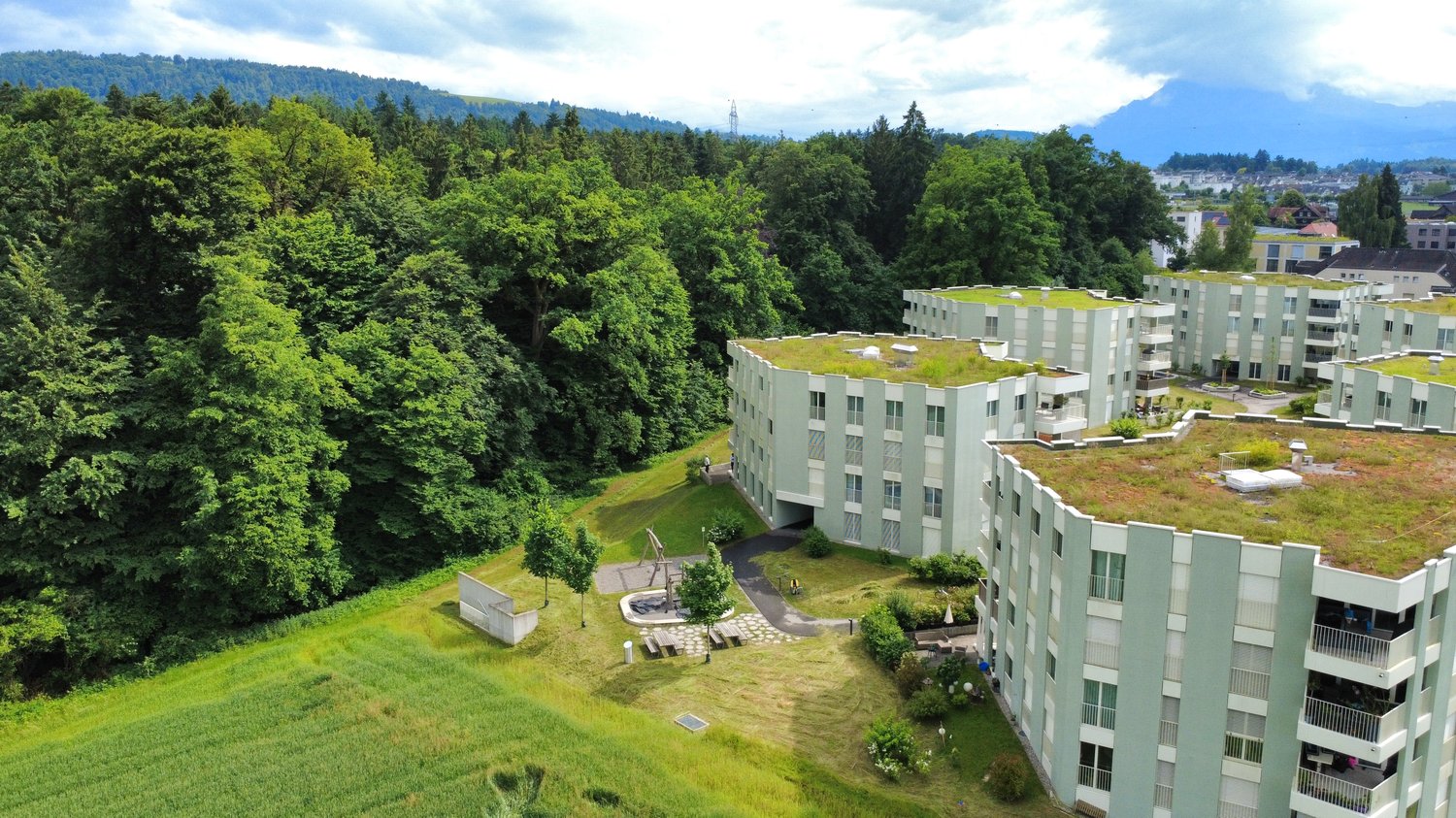 Multi-story apartment building with green roofs, surrounded by lush green trees and vegetation, with parking spaces visible on the ground level