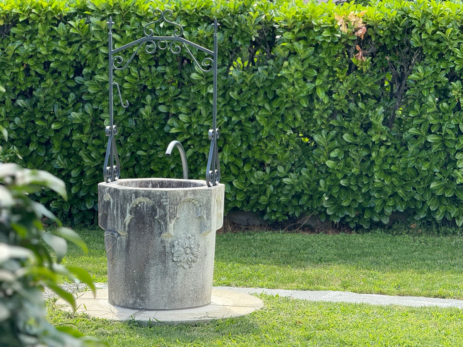 concrete water well with a metal pump, hedge, green lawn, gravel pathway
