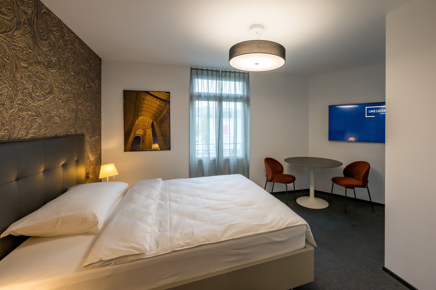 Spacious bedroom with a large bed, leather headboard, and a sitting area with a round table and two chairs. The room has large windows with curtains, and a flat-screen TV on the wall. The decor includes a framed artwork and a drum-shaped light fixture.