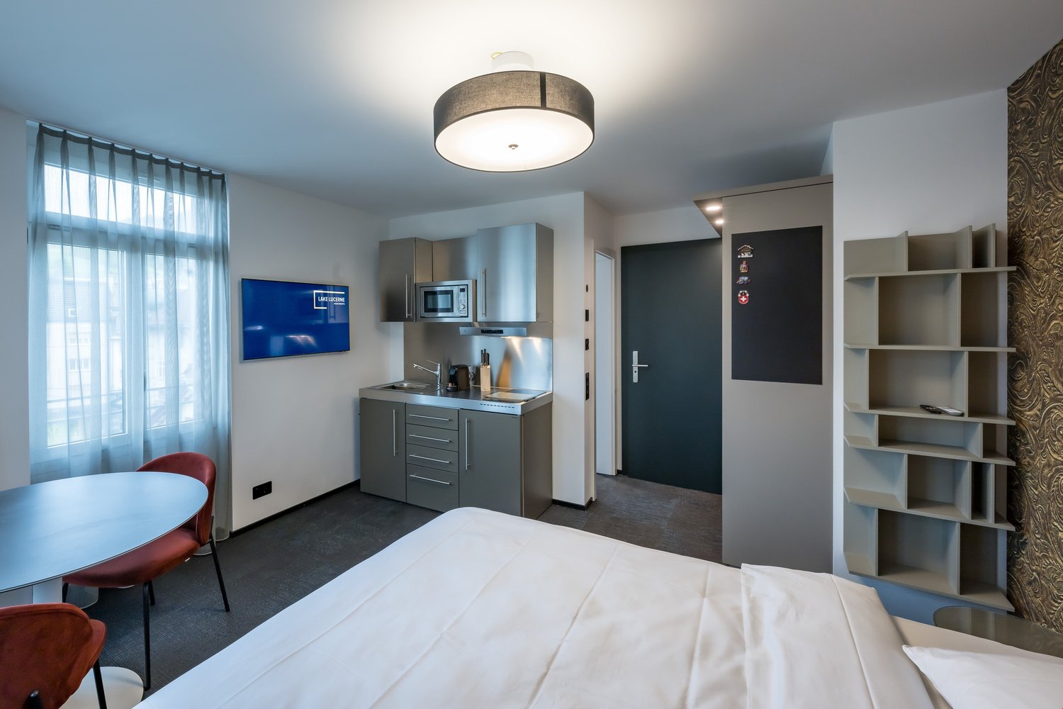 The image shows a modern studio apartment with a kitchenette, a bed, and a living area. The kitchenette has a microwave, a sink, and a dishwasher. The living area has a TV mounted on the wall and a small dining table with chairs. The room has large window