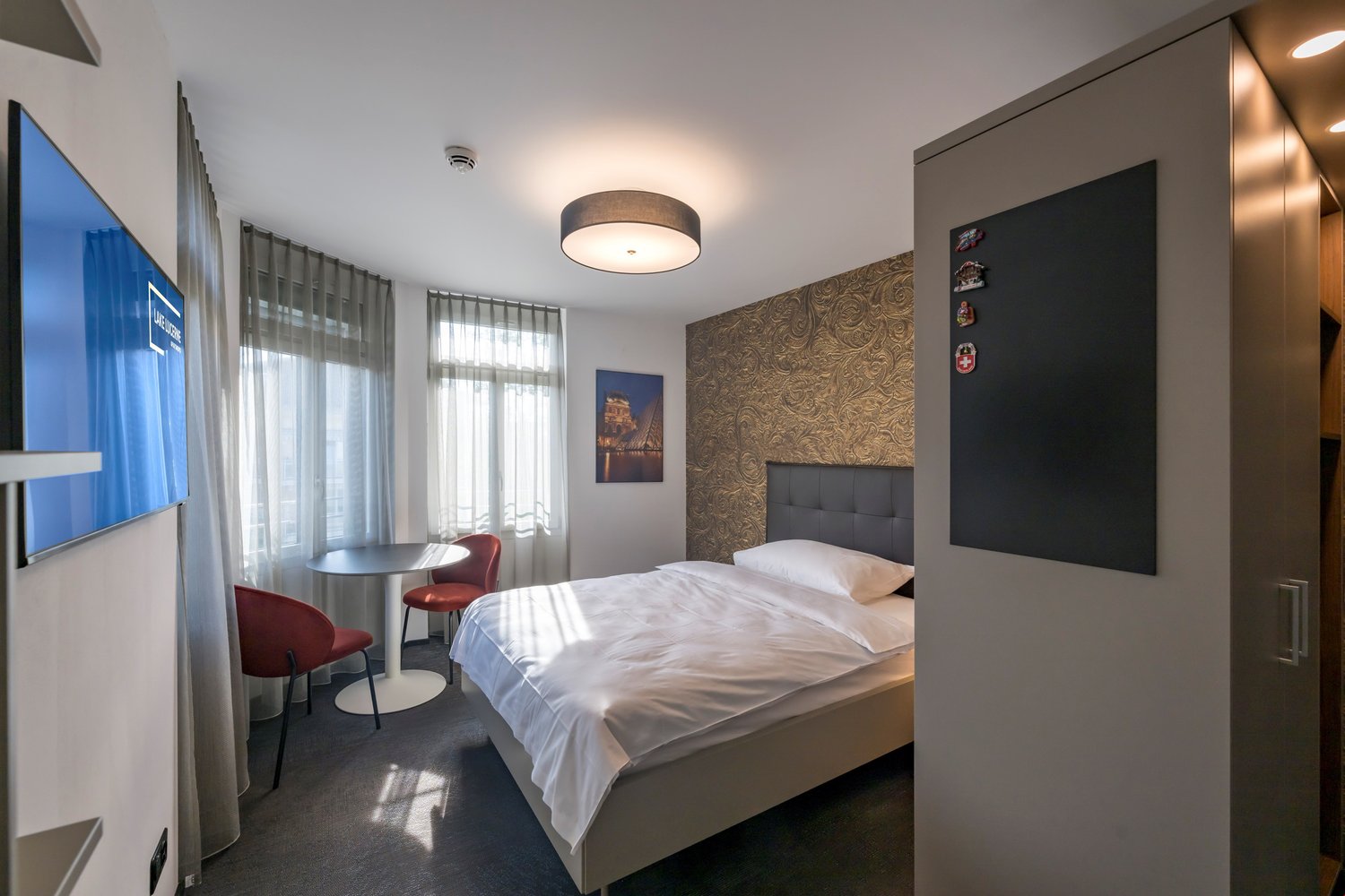 A bedroom with a single bed, bedside tables, a TV, a painting on the wall, and two red chairs next to a round table.