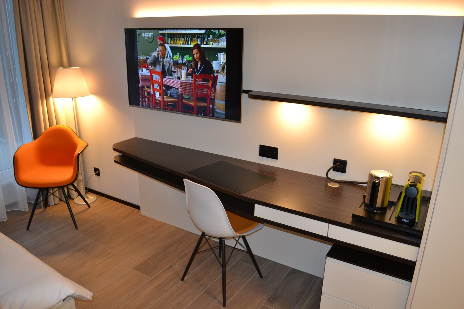 Modern living room with a large TV mounted on the wall, a dark wooden TV stand with drawers, an orange accent chair, and a white chair at a desk area. The room has hardwood flooring and curtains covering a window or balcony door.