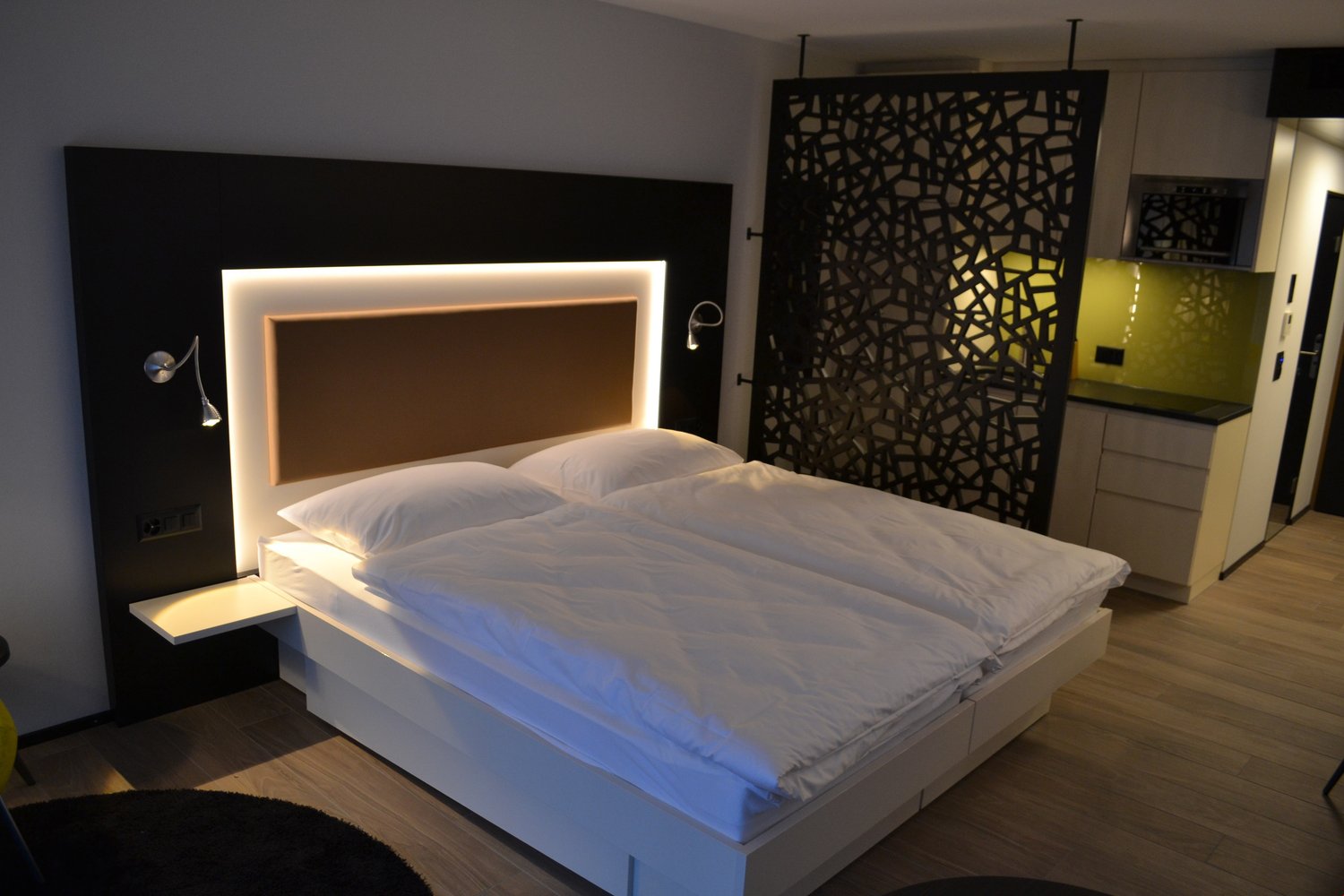 Modern bedroom with a large bed, white bedding, and a backlit headboard. The room also features a decorative screen on the wall and a kitchenette area.