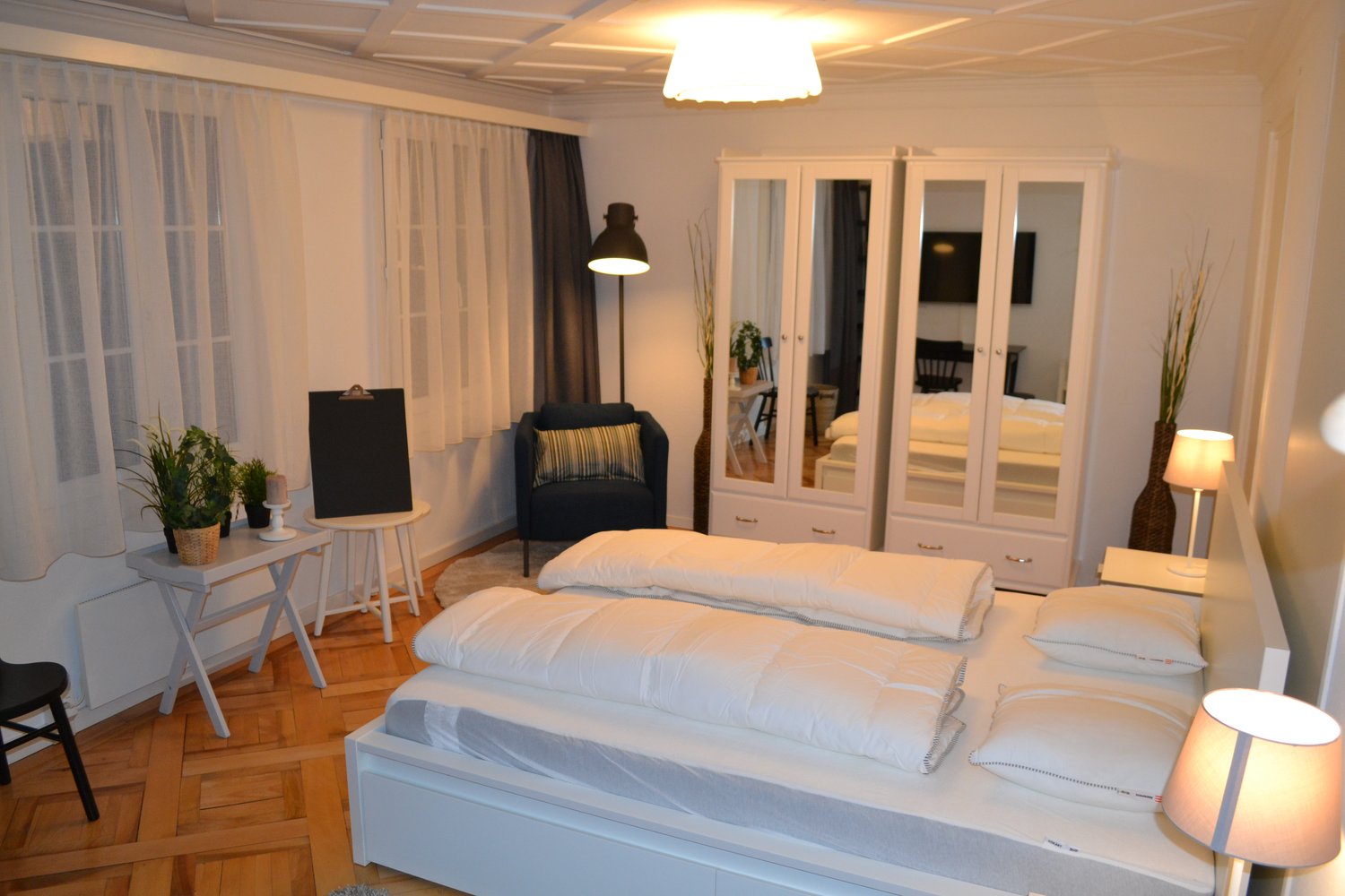 modern bedroom with double bed, white walls, wooden floor, lamps, white furniture, double doors, plants, curtains