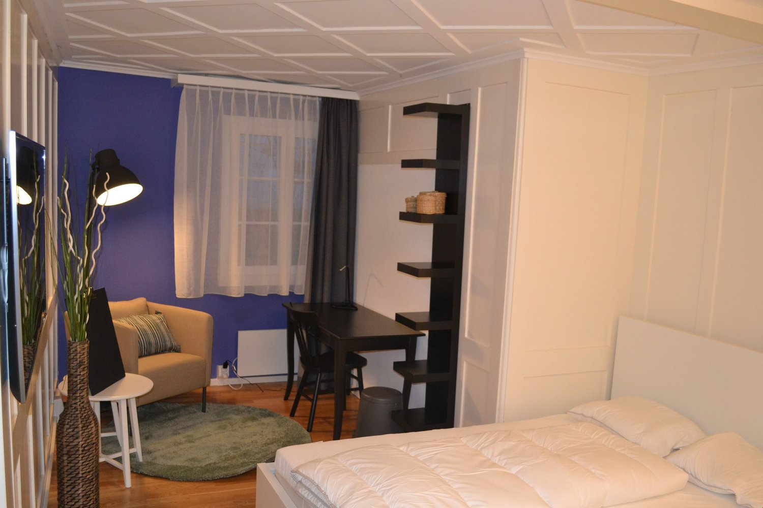 The image shows a studio apartment with a living area, a dining area, and a sleeping area. The living area has a couch and a rug, while the dining area has a small table and chairs. The sleeping area has a bed. There is a window with curtains, and a balco