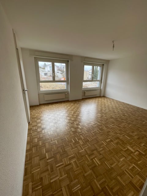 Spacious living room with hardwood parquet flooring, large windows providing natural light, and white walls.