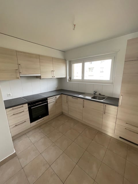 The image shows a modern, L-shaped kitchen with light-colored wood cabinets, black countertops, and a tiled floor. The kitchen appears to have a built-in oven and sink, and there is a window providing natural light.