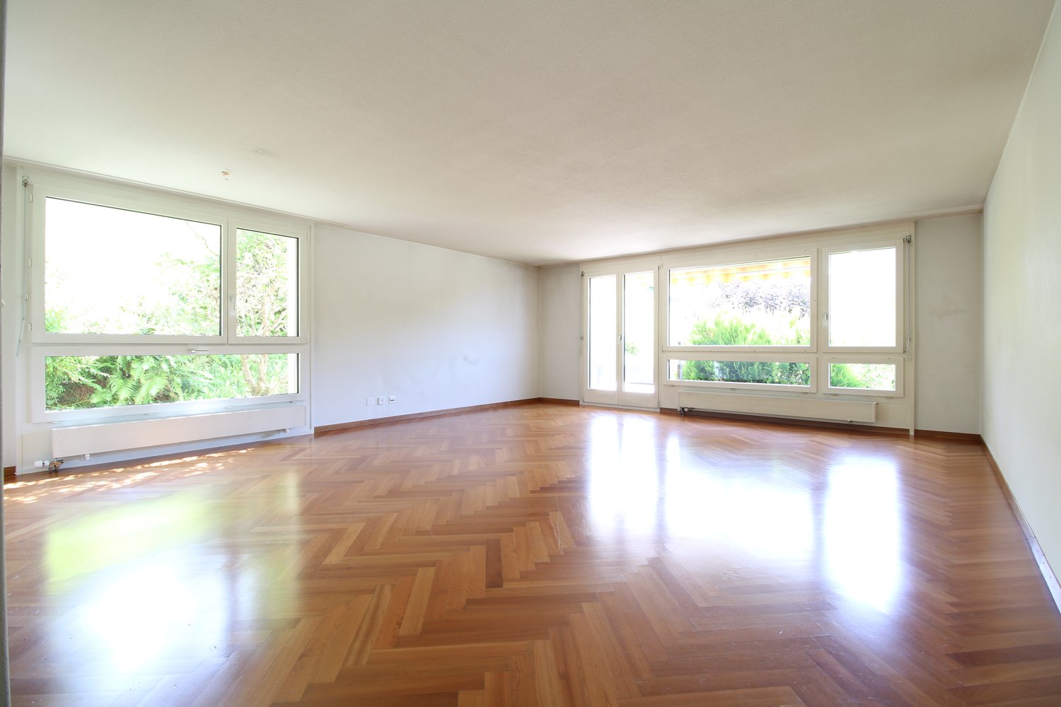 Empty room with wooden parquet flooring, white walls, large windows with sliding doors