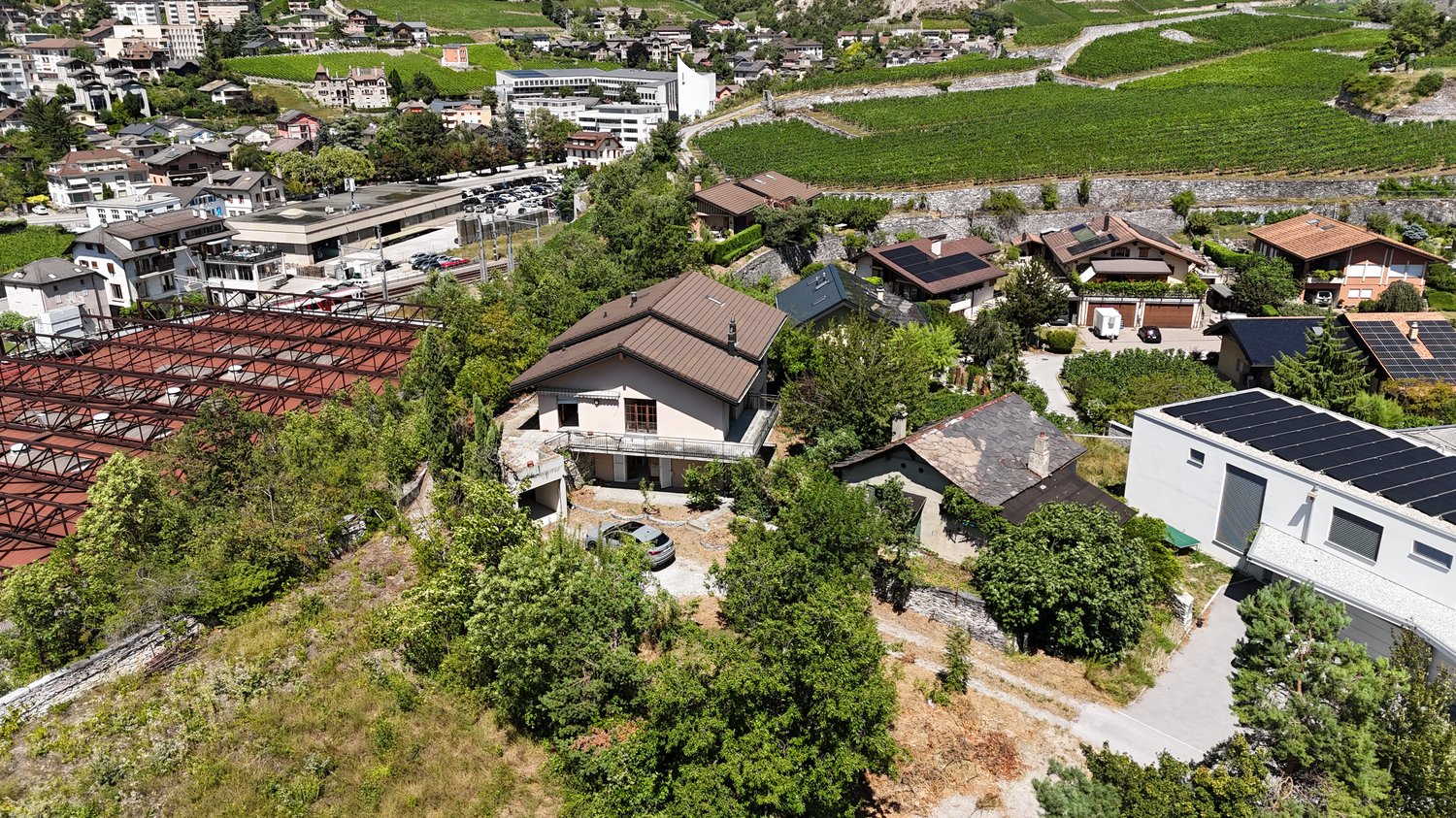 Detached house, brown roof, several windows, parked car, surrounding trees, nearby road, multiple houses in the vicinity, greenery, vineyards in the background, red-roofed structure in the left foreground