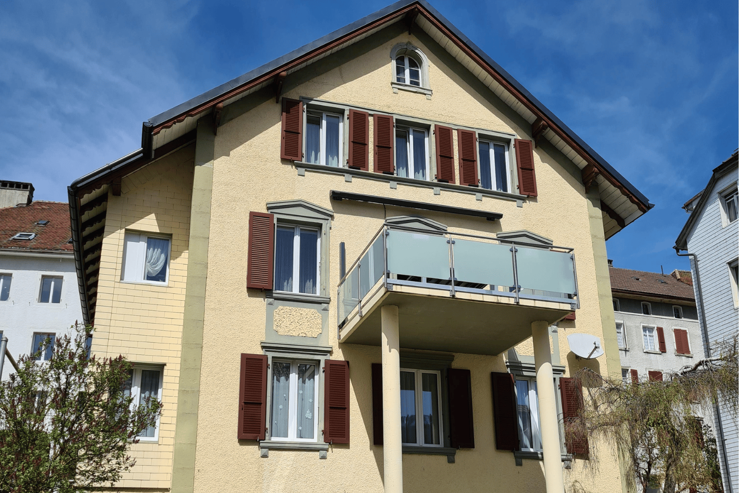 3 story beige house with brown shutters, glass balcony on the second floor, beige pillars, brown roof