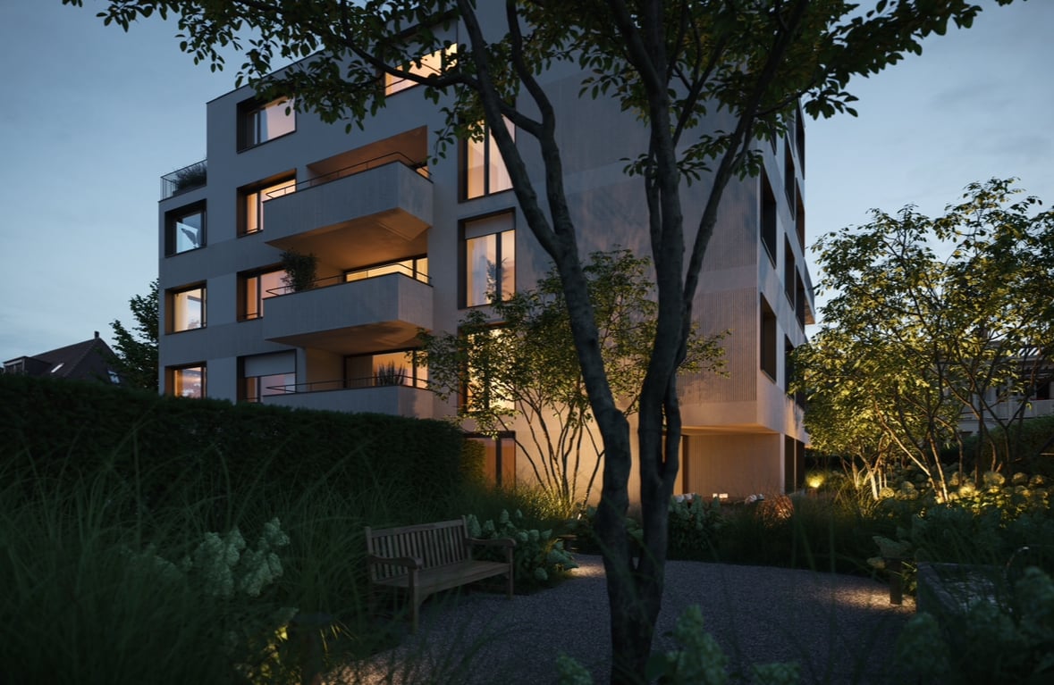 Multi-story apartment building with balconies, surrounded by trees and landscaped garden area with a bench. The building has a modern, minimalist design with large windows and a mix of materials like wood and concrete.