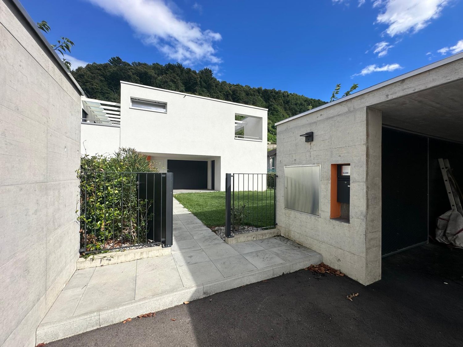modern concrete house, black fence, two entry gates, driveway, green lawn, one covered parking space
