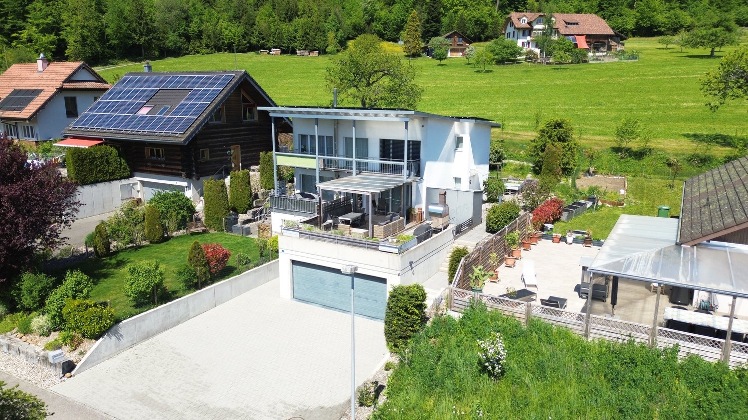 Two-story modern house with solar panels on the roof, surrounded by a lush green garden and trees. The house has a balcony, a garage, and a parking space.