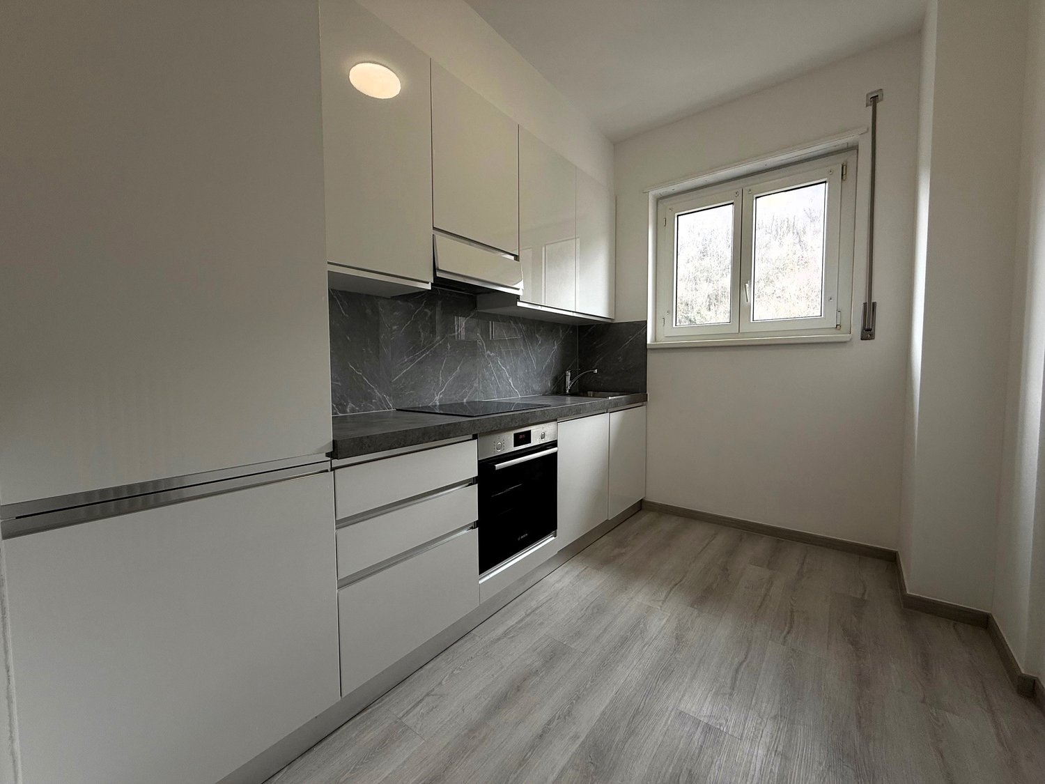 Modern kitchen with white cabinets, wooden flooring, marble countertops, integrated appliances, and a window.