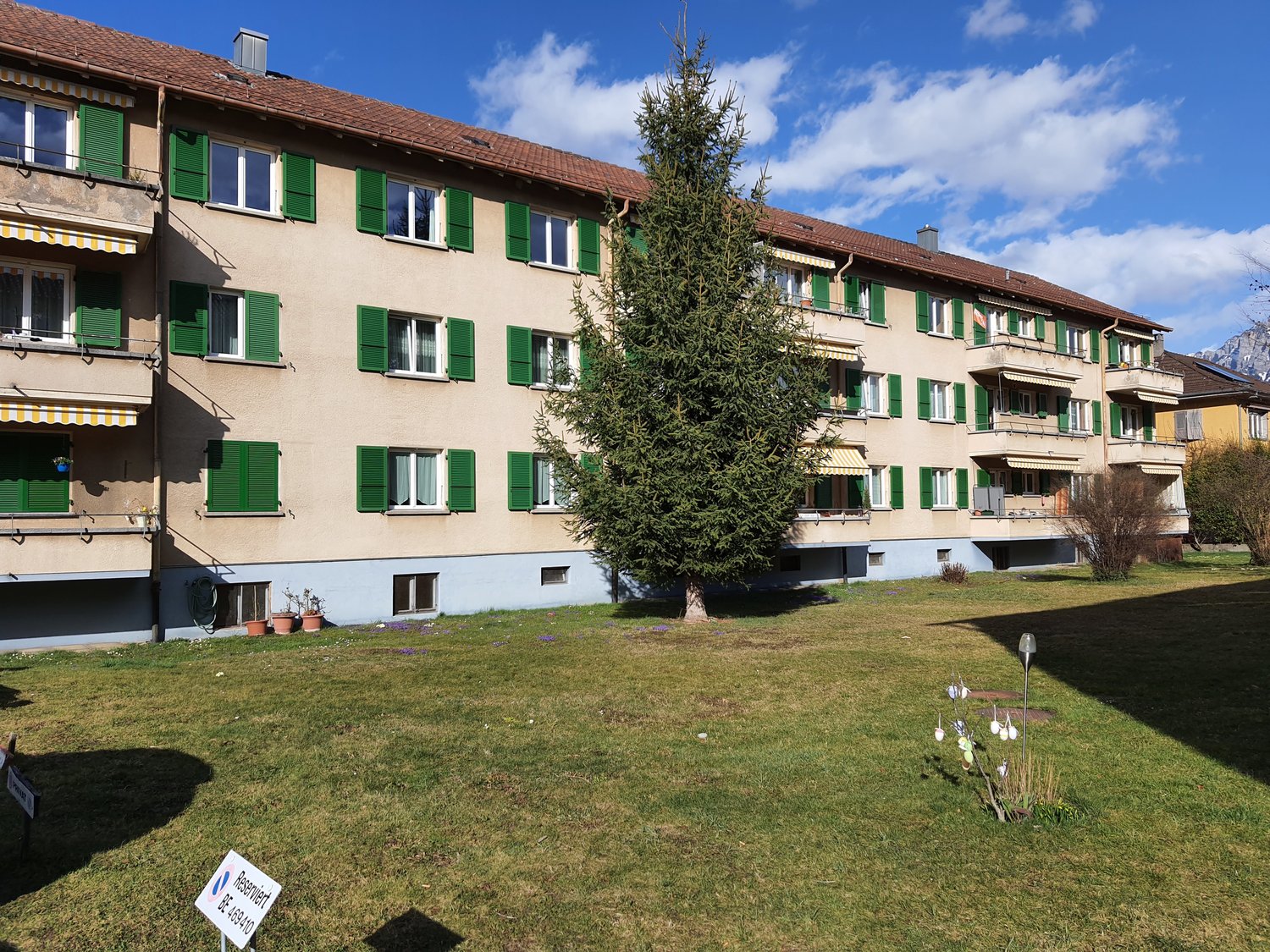 A multi-story apartment building with multiple balconies, green shutters, and a tree in front. There is a well-maintained lawn in front of the building.