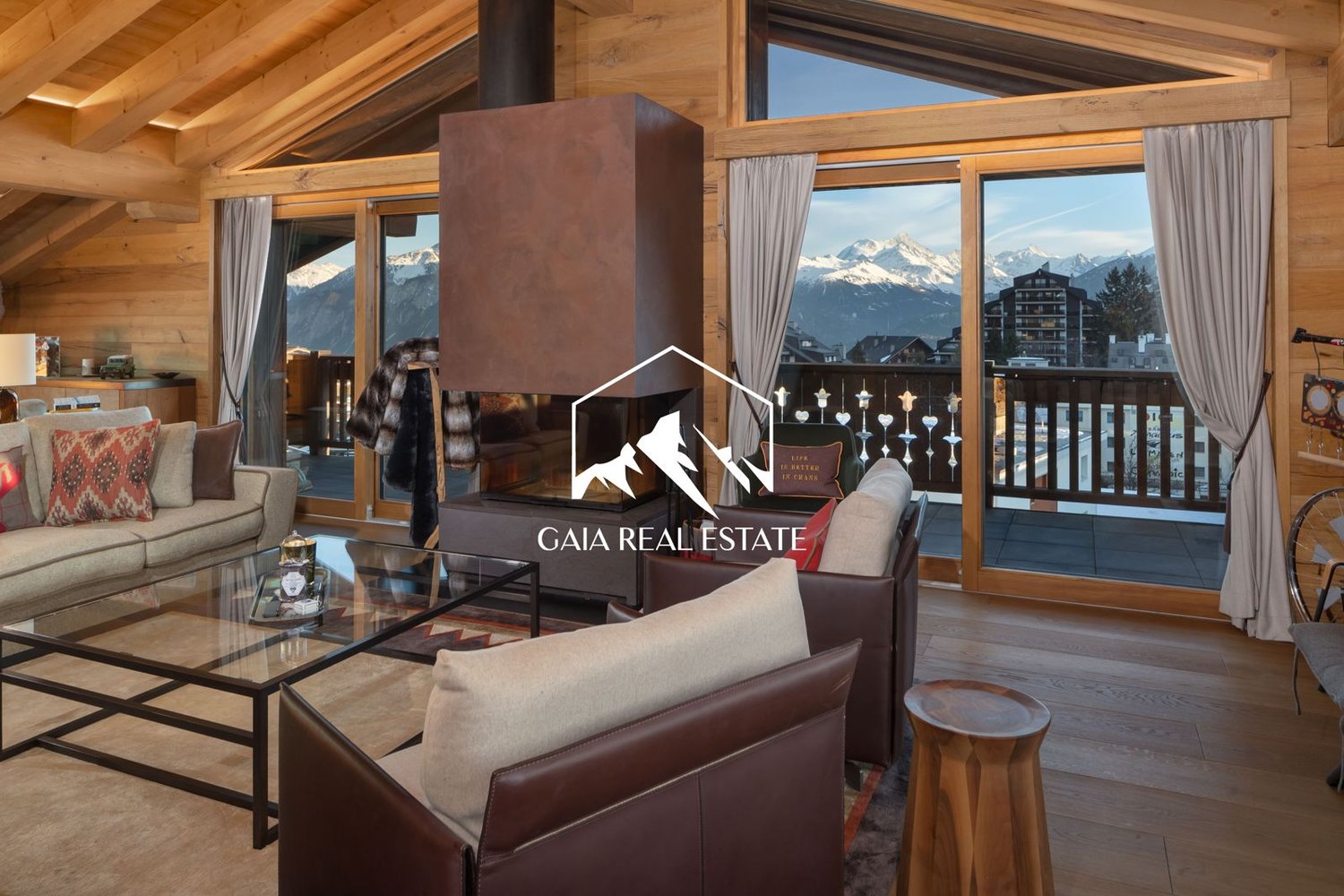 Spacious living room with wooden beams and large windows overlooking snowy mountains, leather sofas, glass coffee table, and a fireplace