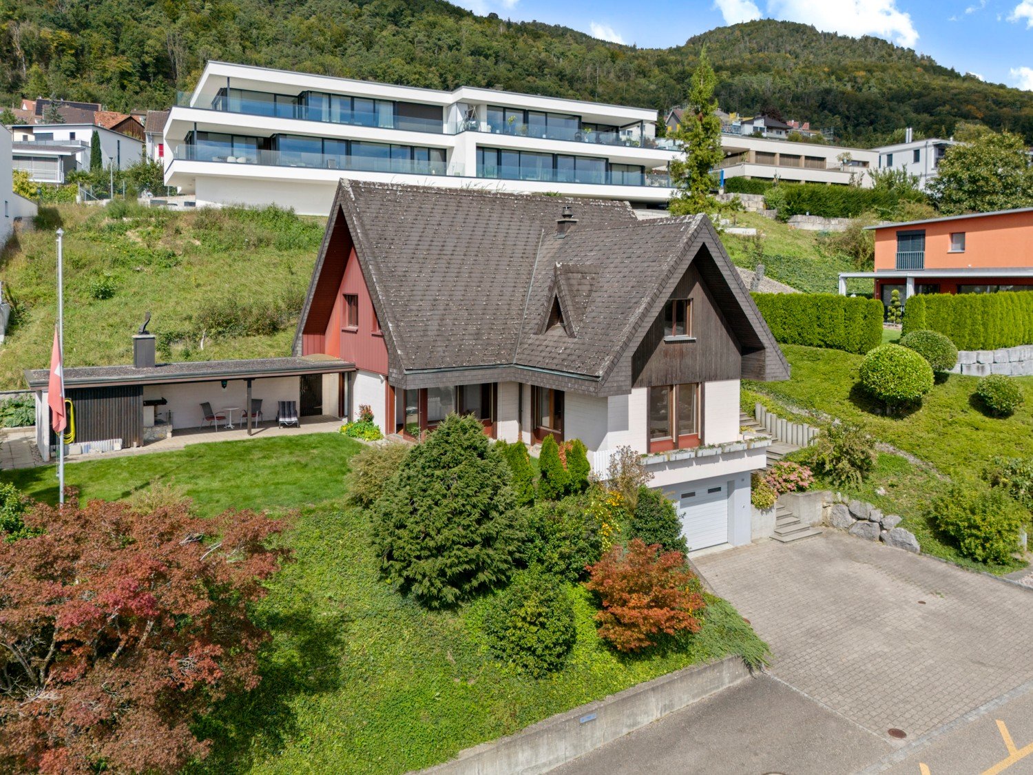 House on slope with garage, driveway, and parking, wooden and red accents, green lawn, and bushes, mountain background.