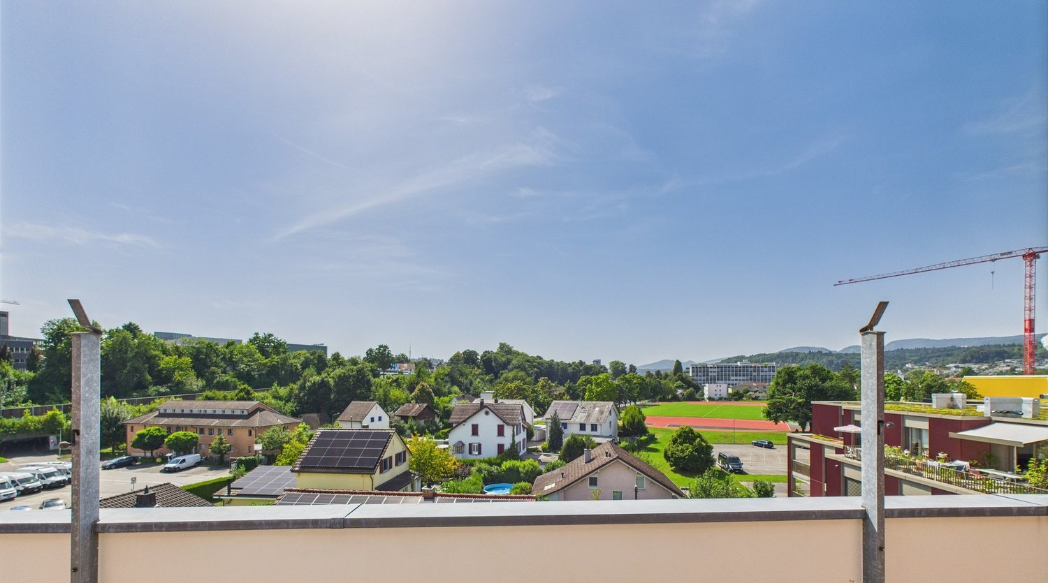 Aerial view of houses, greenery, clear sky, and a crane in the distance, with some parked vehicles.